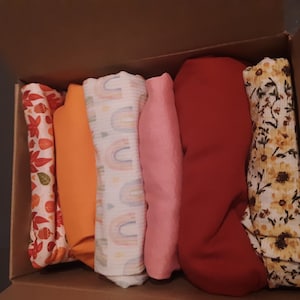 Könnte beinhalten: Eine Schachtel gefüllt mit sechs verschiedenen Stoffproben. Die Proben sind gefaltet und in einer Reihe angeordnet. Die Stoffe sind in verschiedenen Farben und Mustern, darunter Orange, Rosa, Rot, Weiß und ein Blumenmuster.