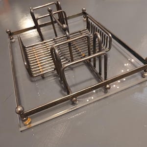 Pode incluir: Um porta-guardanapos de metal vintage com base de vidro transparente. O suporte tem formato retangular com vários compartimentos para guardanapos e uma borda decorativa. O metal tem um acabamento manchado.