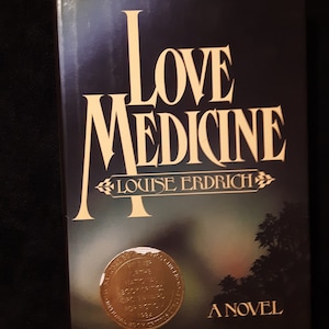 May include: A hardback book titled "Love Medicine" by Louise Erdrich. The book cover is dark blue with gold lettering and a gold seal that reads "Winner of the National Book Critics Circle Award for Fiction 1984". The book is a novel.
