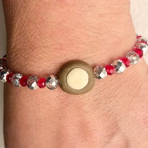 Könnte beinhalten: Ein rotes und silbernes Perlenarmband mit einem braunen und weißen Knopf-Anhänger.