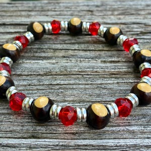 May include: A beaded bracelet featuring alternating dark brown wooden beads with light tan circular insets, faceted translucent red glass beads, and small metallic silver spacers arranged in a repeating pattern around the circular band.