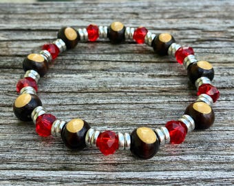 May include: A beaded bracelet featuring alternating dark brown wooden beads with light tan circular insets, faceted translucent red glass beads, and small metallic silver spacers arranged in a repeating pattern around the circular band.