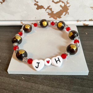 May include: A beaded bracelet with brown wooden beads, red and clear glass beads, and white heart-shaped beads with the letters "J" and "H" and a red heart symbol.