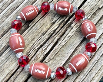 May include: A beaded bracelet featuring alternating football-shaped beads and faceted red beads. The football beads are brown with white stripes and stitching. The bracelet is strung on a clear elastic cord.