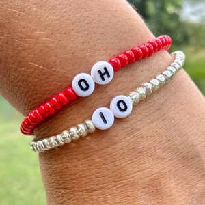 May include: Two beaded bracelets. One is red with white letter beads spelling "OH". The other is silver with white letter beads spelling "IO". The bracelets are worn on a wrist.