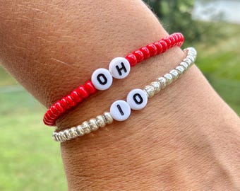May include: Two beaded bracelets. One is red with white letter beads spelling "OH". The other is silver with white letter beads spelling "IO". The bracelets are worn on a wrist.