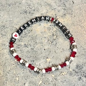 May include: A red and white beaded bracelet with silver spacers and black letter beads spelling "I love Jeremiah Smith".