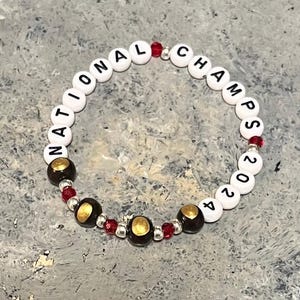 Op de afbeelding: Witte kralen armband met zwarte letters die "NATIONAL CHAMPS 2024" spellen. De armband heeft rode en gouden kralen.