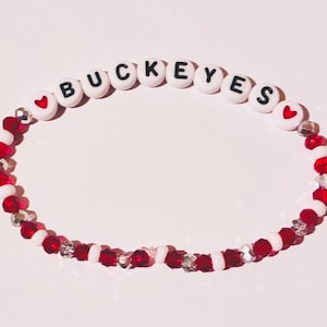 Könnte beinhalten: Ein Perlenarmband mit weißen runden Perlen, die den schwarzen Schriftzug BUCKEYES tragen, flankiert von zwei roten Herzperlen. Der restliche Teil des Armbands besteht aus abwechselnd roten facettierten Kristallperlen, klaren Kristallperlen und kleinen weißen Perlen.