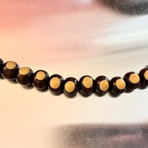 May include: A string of spherical beads arranged in a curved line. Each bead features a dark brown exterior with a circular, light beige or cream-colored inlay centered on the front-facing side, strung together on a thin, clear filament.