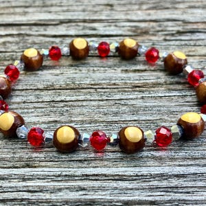 Könnte beinhalten: Ein Perlenarmband mit abwechselnden runden braunen Holzperlen mit hellbeigen Zentren, leuchtend roten facettierten Kristallperlen und kleinen transparenten Zwischenperlen, die in einem sich wiederholenden Muster angeordnet sind.