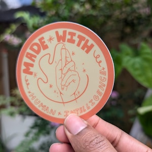 May include: A round sticker with an orange border and a light yellow background. The sticker features a hand drawing a line with a pencil, surrounded by stars. The text "MADE WITH HUMAN + INTELLIGENCE" is written in a circular pattern around the hand.
