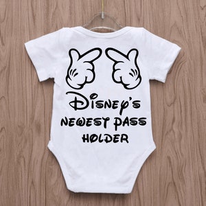May include: White baby onesie with black graphic of Mickey Mouse hands and the text "Disney's Newest Pass Holder." The onesie has short sleeves and is hanging on a clear hanger against a wooden background.