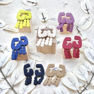 Pode incluir: Seis pares de brincos pendentes com formas geométricas e vários pingentes em forma de lágrima. Os brincos são exibidos em blocos de madeira e estão disponíveis em uma variedade de cores, incluindo verde, roxo, branco, azul, vermelho e marrom.