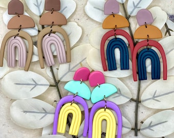 Könnte beinhalten: Drei Paar bunte Ohrringe aus Polymer Clay. Jedes Paar hat ein Regenbogenbogen-Design mit einem Halbkreis und einem kleinen Kreis oben. Die Farben sind Pink, Braun, Türkis, Gelb, Lila und Rot.