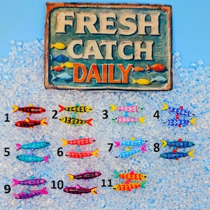 Könnte beinhalten: Ein rustikales Schild mit der Aufschrift "FRESH CATCH DAILY" über einer Sammlung farbenfroher fischförmiger Verzierungen. Die Fische sind in verschiedenen Farben gehalten, darunter Rot, Gelb, Blau und Lila. Das Schild hat ein verwittertes Aussehen.