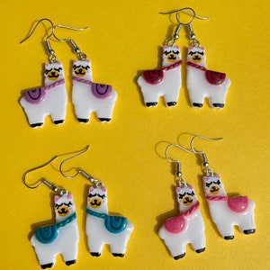 May include: Four pairs of llama earrings. Each pair features a white llama with a different colored saddle. The saddles are purple, red, teal, and pink.