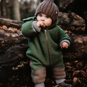 Peut inclure: Un tout-petit portant une combinaison en polaire verte et marron avec un bonnet en laine marron. L'enfant est debout dans un environnement forestier.