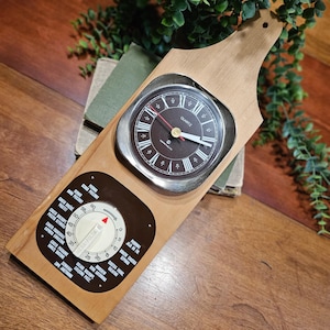 May include: A wall-mounted kitchen clock and timer combination on a light wood board. The upper section features a round quartz clock with a dark brown face and white Roman numerals. Below, a circular timer dial includes Polish text labels.