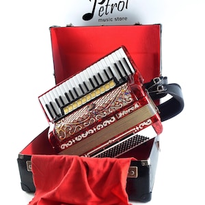 Pode incluir: Um acordeão vermelho e dourado com teclas brancas repousa em um estojo preto aberto com interior vermelho. O acordeão tem detalhes ornamentados e o nome "Horch" é visível. Um pano vermelho está drapeado no estojo. O texto "Petrof music store" está no topo.