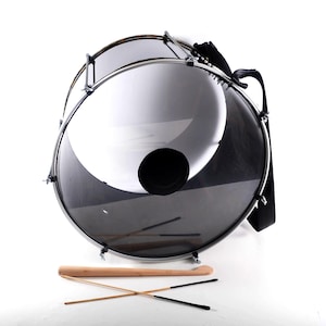May include: A silver-coloured drum with a black strap and two wooden sticks. The drum has a black circular patch on the front.