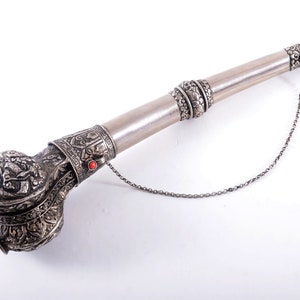 May include: A silver smoking pipe with intricate carvings and a chain attached. The pipe has a bulbous mouthpiece and a long, slender stem. The stem is decorated with a band of silver.