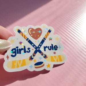 May include: A holographic sticker with the words "girls rule" and hockey-themed graphics. The design features crossed hockey sticks, a heart, a puck, and stars. The sticker has a white border and is held against a pink background.
