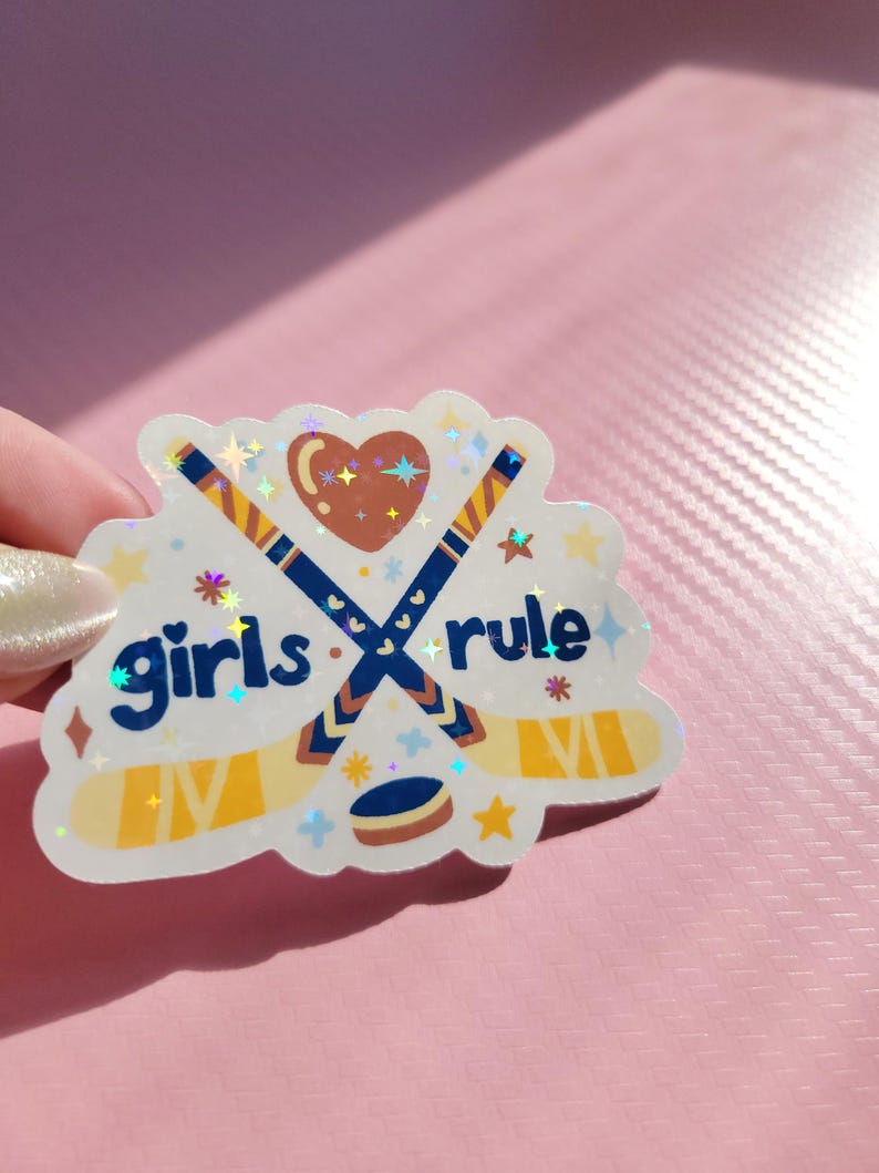 May include: A holographic sticker with the words "girls rule" and hockey-themed graphics. The design features crossed hockey sticks, a heart, a puck, and stars. The sticker has a white border and is held against a pink background.