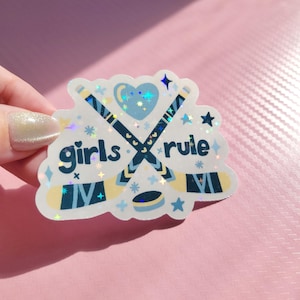 May include: A holographic sticker with the words "girls rule" in a playful font. The sticker features crossed hockey sticks, a heart, stars, and a hockey puck in shades of blue and white. The sticker is held against a pink background.