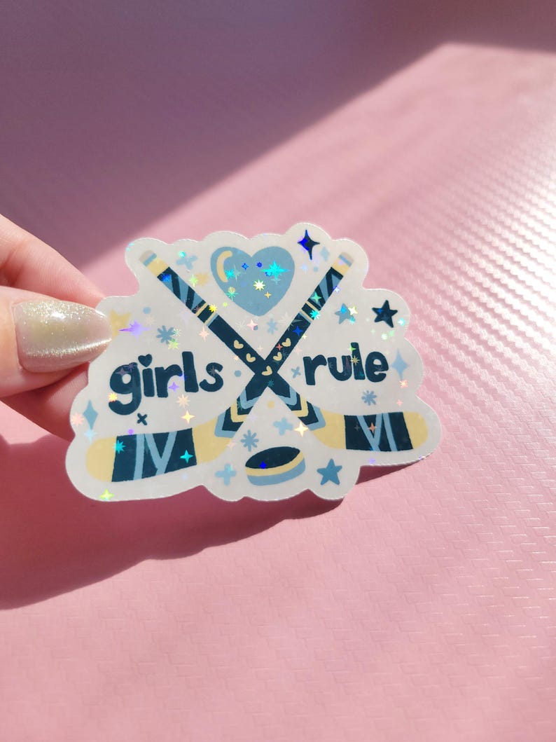 May include: A holographic sticker with the words "girls rule" in a playful font. The sticker features crossed hockey sticks, a heart, stars, and a hockey puck in shades of blue and white. The sticker is held against a pink background.