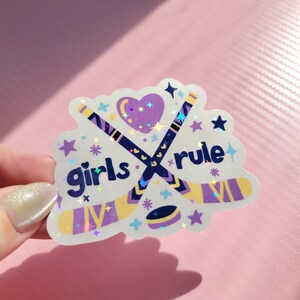 May include: A holographic sticker with the words "girls rule" and hockey sticks. The sticker features a purple heart, stars, and a hockey puck. The hockey sticks are navy blue and yellow. The sticker is held by a hand.
