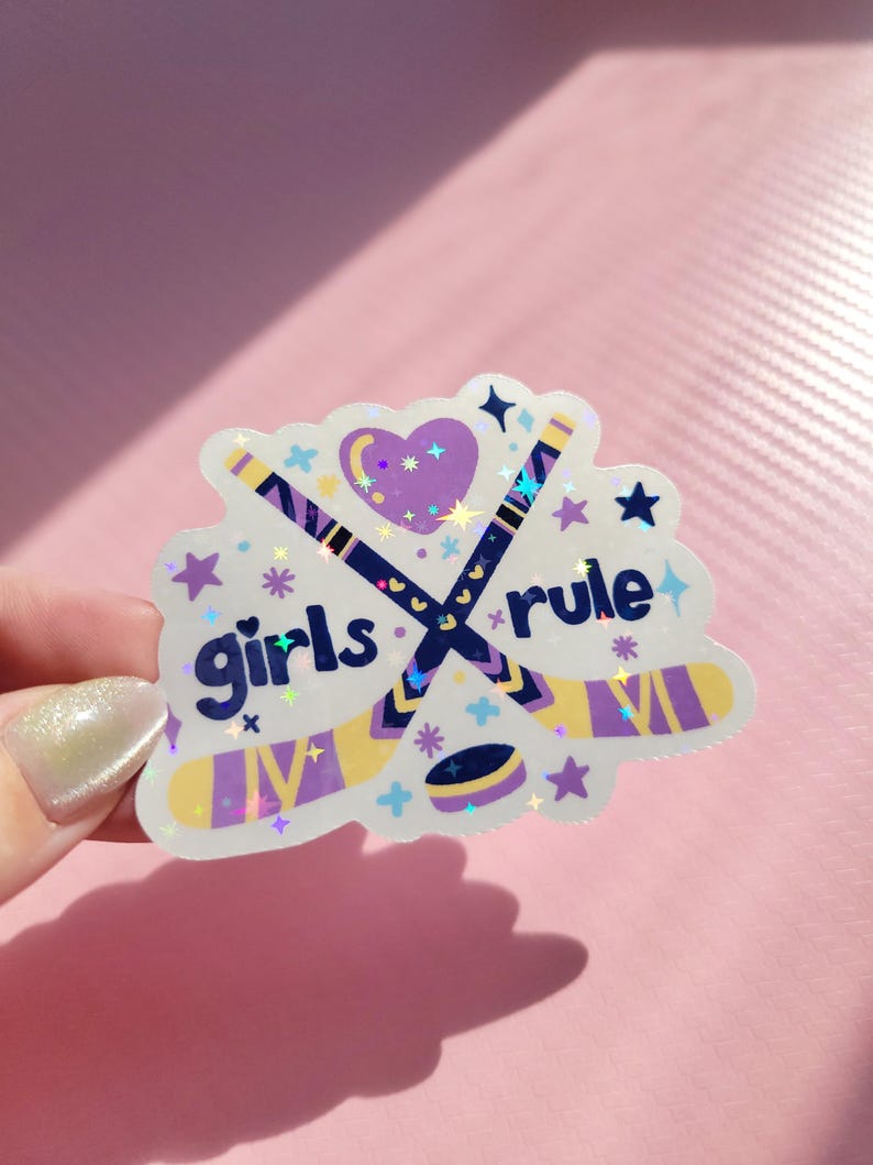 May include: A holographic sticker with the words "girls rule" and hockey sticks. The sticker features a purple heart, stars, and a hockey puck. The hockey sticks are navy blue and yellow. The sticker is held by a hand.
