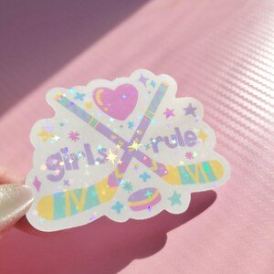 May include: A holographic sticker with the text "Girls Rule" and hockey-themed graphics. The sticker features crossed hockey sticks, a puck, a heart, and stars in pastel colors. The sticker is on a pink background.