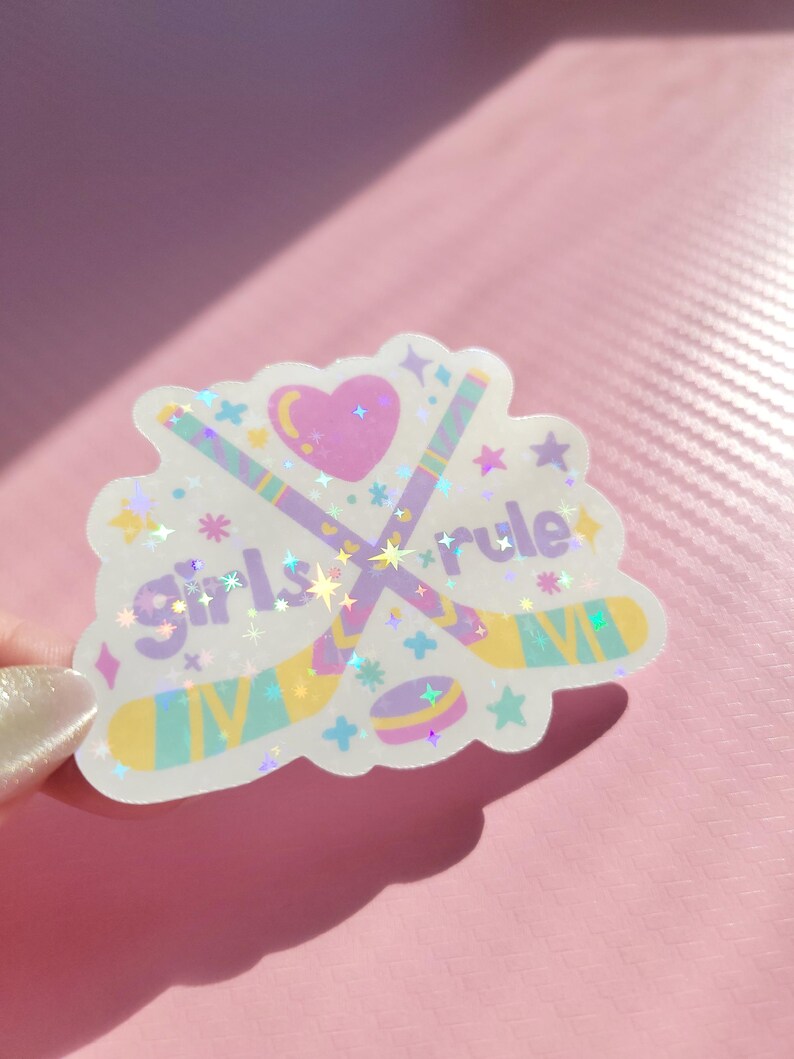 May include: A holographic sticker with the text "Girls Rule" and hockey-themed graphics. The sticker features crossed hockey sticks, a puck, a heart, and stars in pastel colors. The sticker is on a pink background.