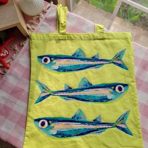 Op de afbeelding: Een limoengroene draagtas met een herhalend patroon van drie gestileerde vissen. Elke vis is afgebeeld in tinten blauw, groenblauw en wit met opvallende ronde ogen. De tas heeft twee bijpassende limoengroene stoffen handvatten aan de bovenkant.