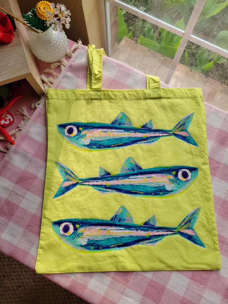 Op de afbeelding: Een limoengroene draagtas met een herhalend patroon van drie gestileerde vissen. Elke vis is afgebeeld in tinten blauw, groenblauw en wit met opvallende ronde ogen. De tas heeft twee bijpassende limoengroene stoffen handvatten aan de bovenkant.