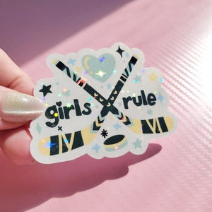 May include: A holographic sticker with the words "girls rule" and crossed hockey sticks. The sticker features a heart, stars, and other decorative elements in pastel colors. The sticker is held by a hand.
