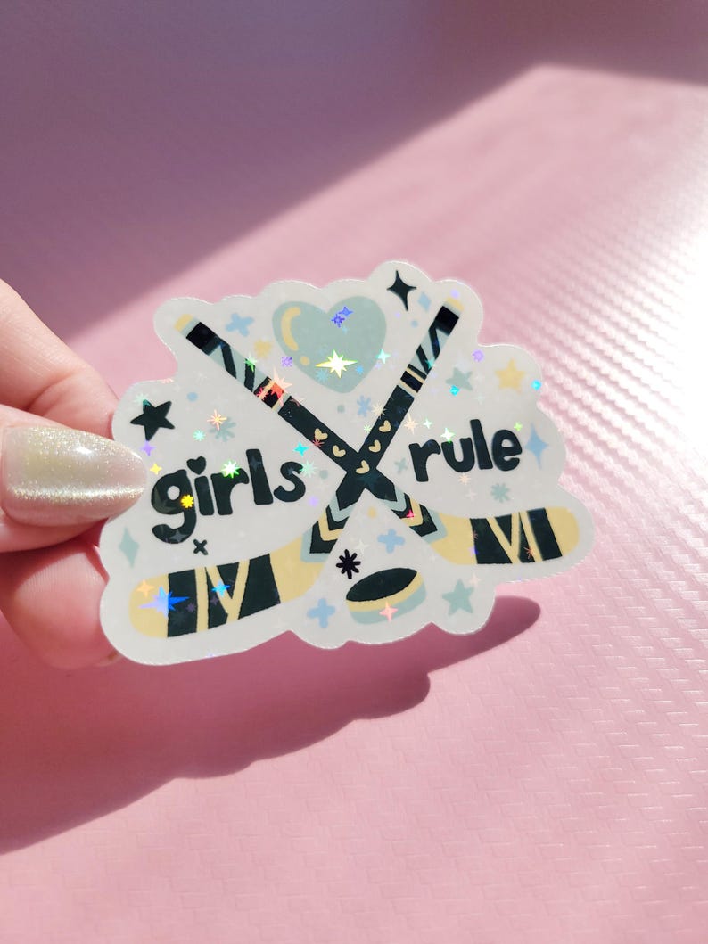 May include: A holographic sticker with the words "girls rule" and crossed hockey sticks. The sticker features a heart, stars, and other decorative elements in pastel colors. The sticker is held by a hand.