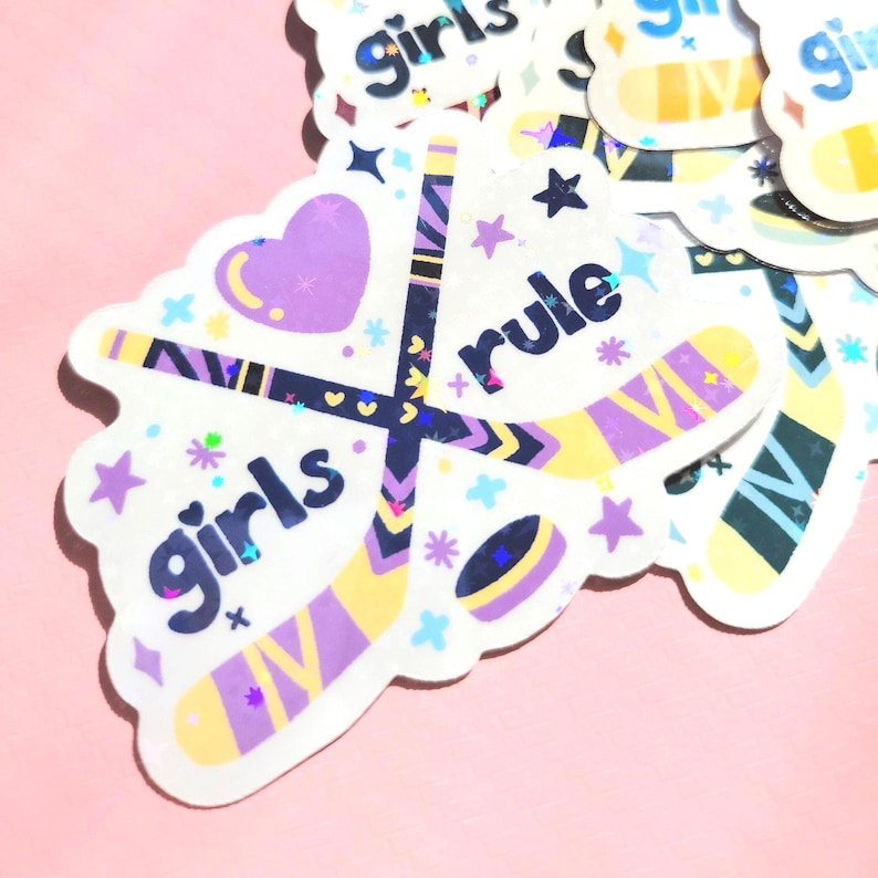 May include: A collection of stickers with a hockey theme. The stickers feature hockey sticks, a puck, and the words "girls rule." The design incorporates purple, yellow, and blue colors with star and heart accents. The stickers have a holographic effect.