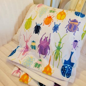 May include: White blanket featuring a colorful pattern of various insects in shades of orange, green, blue, purple, and yellow. The blanket is folded and appears soft, with a plush texture. The design includes beetles and other bugs.