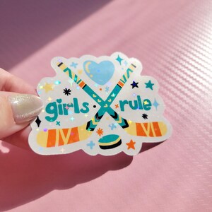 May include: A holographic sticker with the text "girls rule" and hockey-themed graphics. The design features crossed hockey sticks, a puck, stars, and a heart. The sticker has a white background and is held against a pink surface.