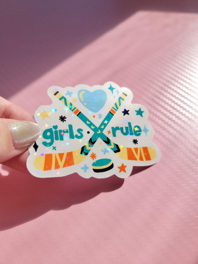 May include: A holographic sticker with the text "girls rule" and hockey-themed graphics. The design features crossed hockey sticks, a puck, stars, and a heart. The sticker has a white background and is held against a pink surface.
