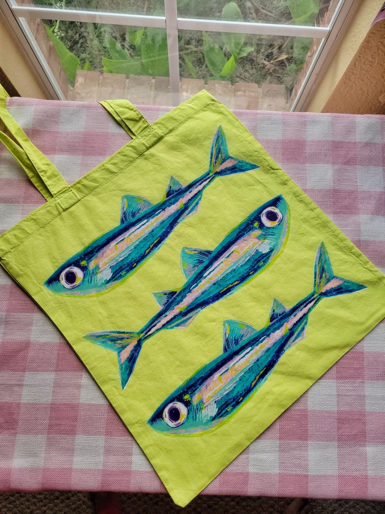 Op de afbeelding: Een limoengroene draagtas met een handgeschilderd ontwerp van drie vissen die diagonaal zijn gerangschikt. De vissen zijn uitgevoerd in tinten blauw, groenblauw en wit met opvallende ronde ogen en zichtbare penseelstreektexturen op hun lichamen.