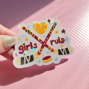 May include: A holographic sticker with the words "girls rule" in red, featuring crossed hockey sticks, a heart, and a hockey puck. The sticker has a white border with star and flower accents, and is held by a hand.