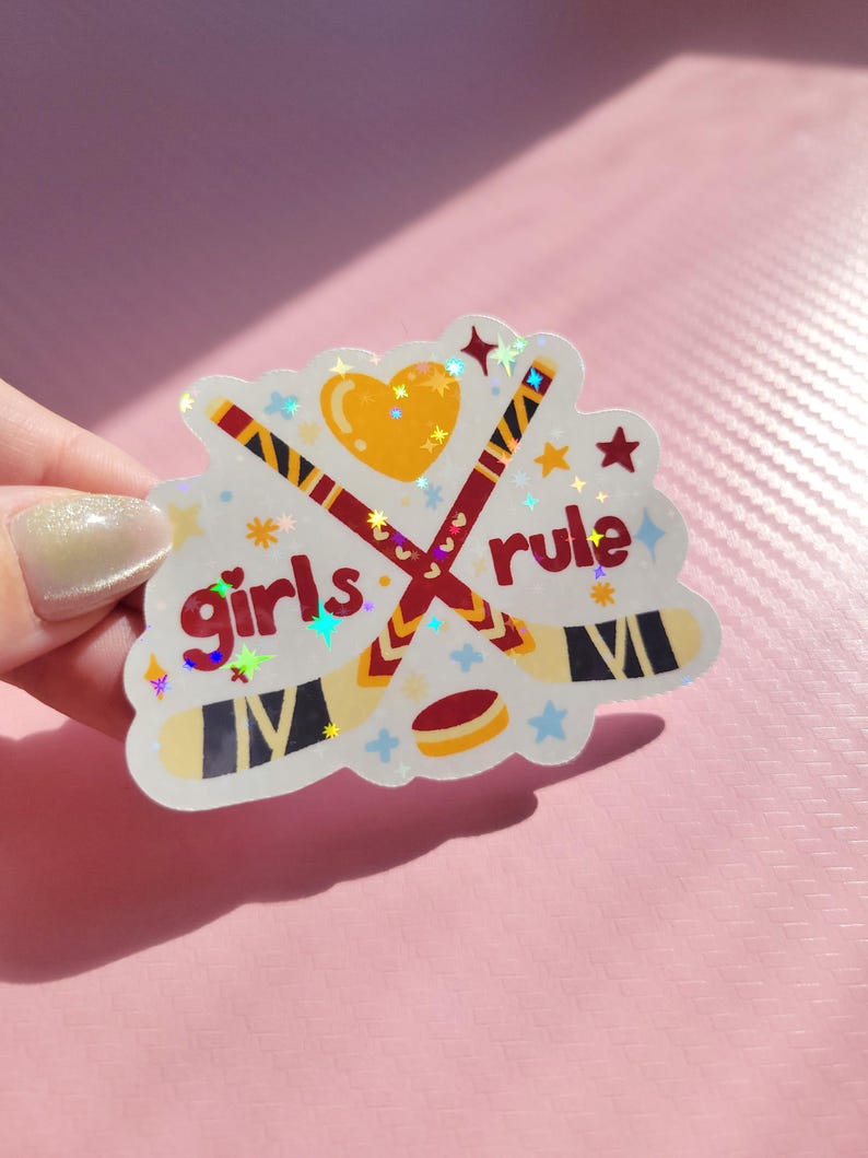 May include: A holographic sticker with the words "girls rule" in red, featuring crossed hockey sticks, a heart, and a hockey puck. The sticker has a white border with star and flower accents, and is held by a hand.
