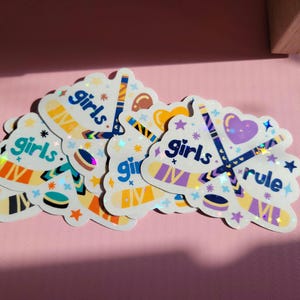 May include: A collection of holographic stickers with a hockey theme. Each sticker features hockey sticks, pucks, and the words "girls" and "rule" in a playful font. The stickers have a colorful design with purple, blue, orange, and yellow accents.