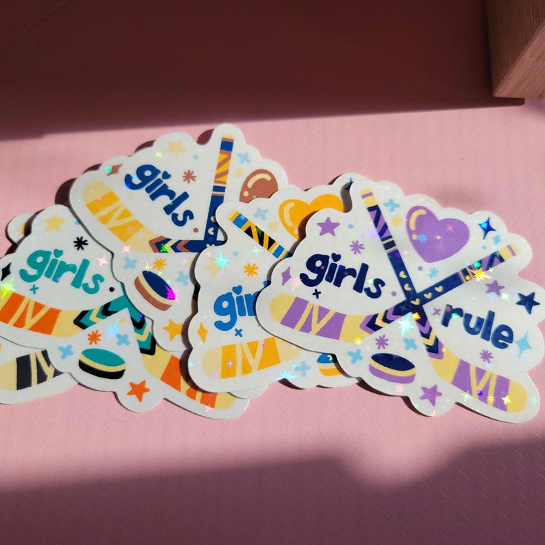 May include: A collection of holographic stickers with a hockey theme. Each sticker features hockey sticks, pucks, and the words "girls" and "rule" in a playful font. The stickers have a colorful design with purple, blue, orange, and yellow accents.