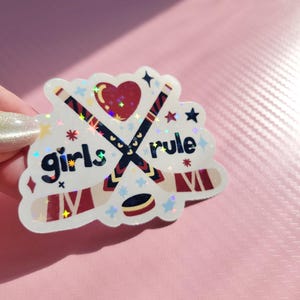 May include: A holographic sticker with the words "girls rule" and hockey-themed graphics. The design features crossed hockey sticks, a heart, stars, and a hockey puck. The sticker has a white border and is on a pink background.