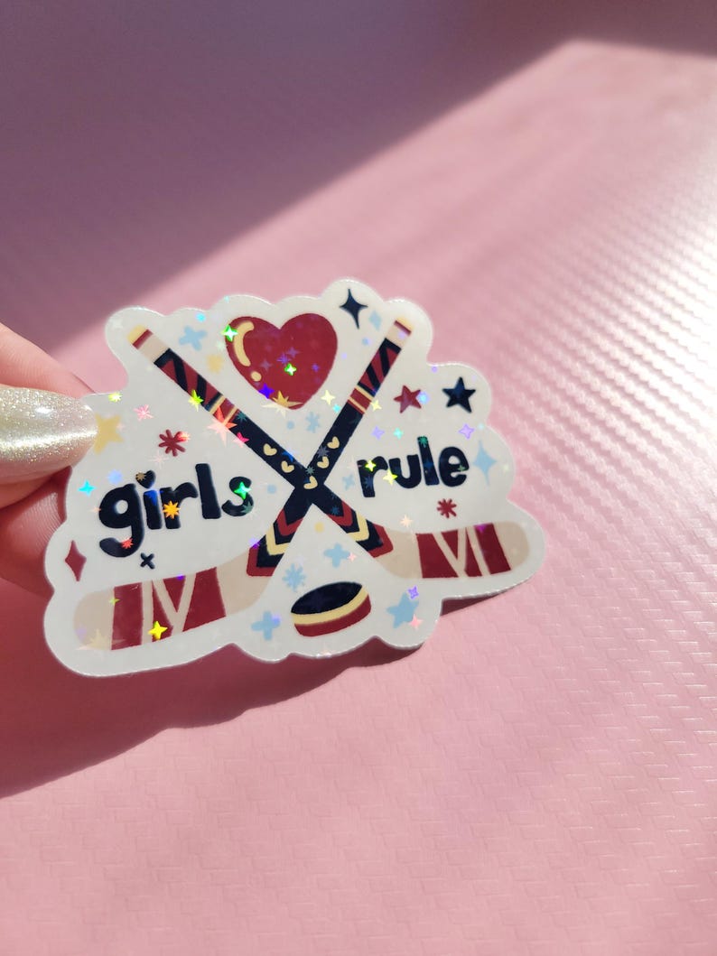May include: A holographic sticker with the words "girls rule" and hockey-themed graphics. The design features crossed hockey sticks, a heart, stars, and a hockey puck. The sticker has a white border and is on a pink background.