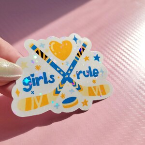 May include: A holographic sticker with the words "Girls rule" and hockey-themed graphics. The design features crossed hockey sticks, a puck, a heart, and stars in blue, yellow, and white. The sticker has a glittery, iridescent finish.